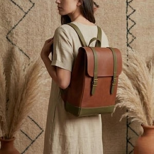 Puede incluir: Una mochila de cuero marrón con detalles en verde oliva. La mochila tiene correas ajustables y un cierre de solapa con dos hebillas. La base y las correas son de color verde oliva. La mochila se está usando.