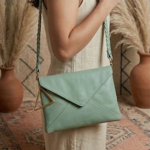 May include: A light green leather crossbody bag with a braided strap. The bag has a triangular gold-colored accent on the front. The bag is worn over the shoulder.
