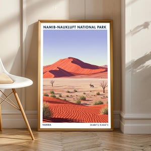 May include: Framed art print of Namib-Naukluft National Park in Namibia. The artwork shows a desert landscape with red sand dunes, sparse vegetation, and a blue sky. The text "NAMIB-NAUKLUFT NATIONAL PARK" is at the top, and "NAMIBIA" and coordinates are at the bottom.