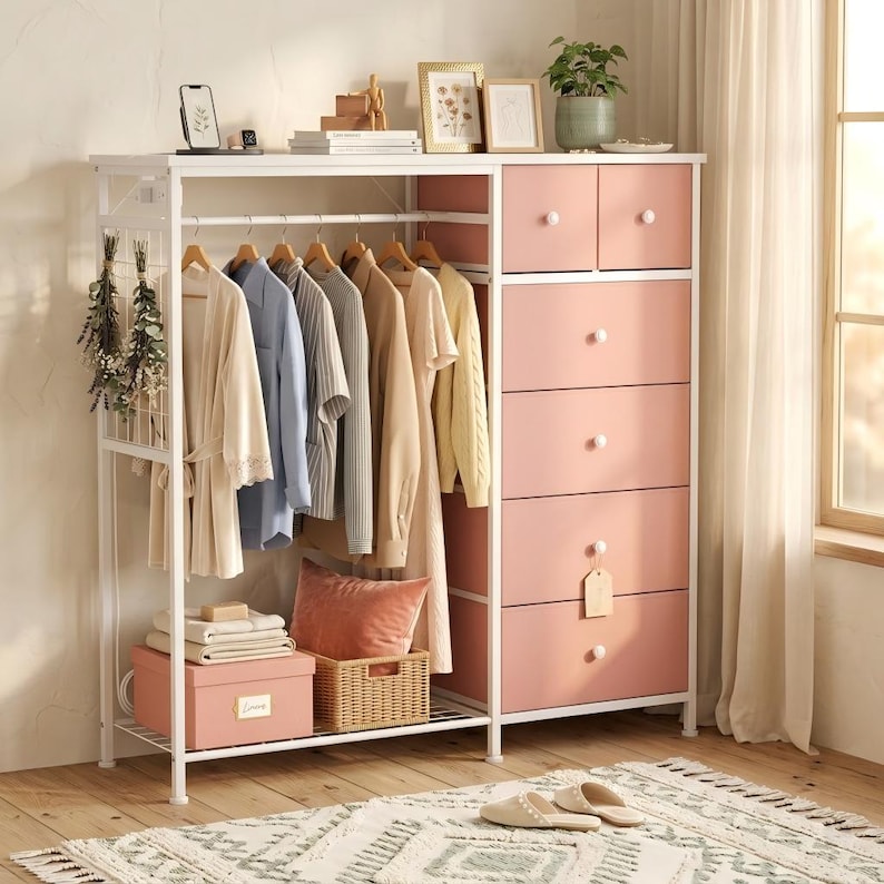 May include: A white metal clothing rack with a pink dresser. The rack holds various garments on wooden hangers. A pink box and a woven basket sit on a lower shelf. The dresser has six drawers with white knobs. A rug and slippers are in the foreground.
