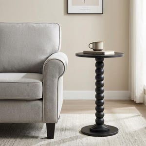 May include: A black side table with a round top and a central pedestal base featuring a stacked sphere design. A beige ceramic mug sits on top of a closed book placed on the table surface. The table has a smooth finish and a circular base.