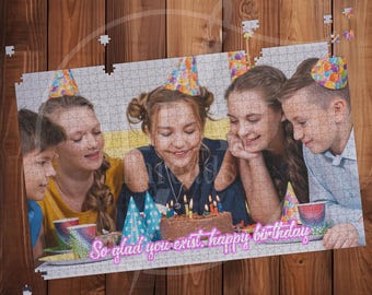 May include: A jigsaw puzzle featuring a group of children celebrating a birthday. The puzzle shows a chocolate cake with lit candles, party hats, and the text "So glad you exist, happy birthday". The puzzle is set against a wooden background.