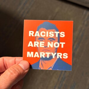 May include: Square sticker with a red background and the white text "RACISTS ARE NOT MARTYRS". The sticker features a portrait of a man in a blue suit. The sticker is held by a hand.