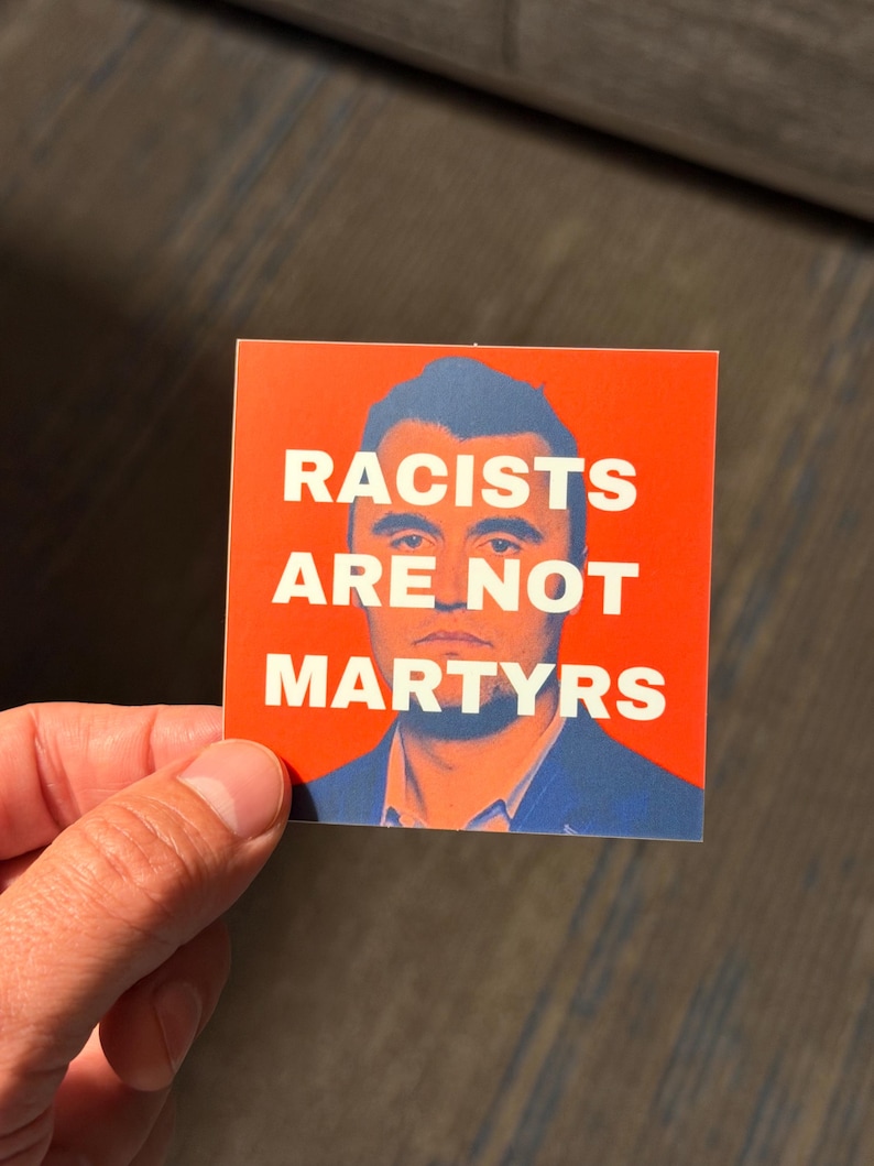 May include: Square sticker with a red background and the white text "RACISTS ARE NOT MARTYRS". The sticker features a portrait of a man in a blue suit. The sticker is held by a hand.