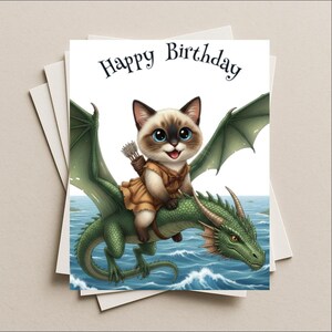 May include: A birthday card featuring a cartoon cat riding a green dragon over blue water. The cat has blue eyes and is wearing a brown outfit with a quiver of arrows. The words "Happy Birthday" are at the top.