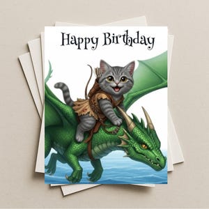May include: A birthday card featuring a gray tabby cat riding a green dragon. The cat wears a brown outfit and a quiver of arrows. The words "Happy Birthday" are written in black script above the illustration. The card is on a stack of white cards.