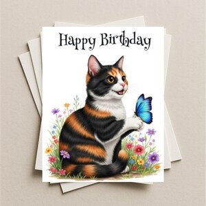 May include: A greeting card with a watercolor illustration of a calico cat holding a blue butterfly. The card reads "Happy Birthday" in black script. Colorful flowers and green grass surround the cat. The card is on a stack of white cards.