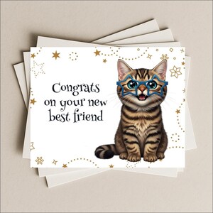 May include: A stack of greeting cards with a cartoon cat wearing blue star-shaped glasses. The cat is brown with black stripes and is smiling. The card reads "Congrats on your new best friend" in black text, with gold star and snowflake accents.