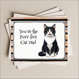 May include: Greeting card featuring a black and white cat with yellow eyes. The card has the text "You're the Purr-fect Cat Dad" on a white background, framed with wood and black accents. Several blank cards are visible.