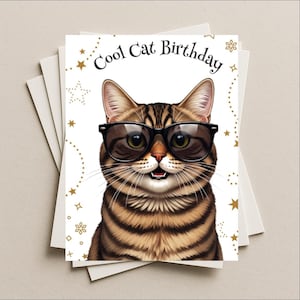 May include: A stack of white greeting cards with a "Cool Cat Birthday" design. The card features a tabby cat wearing black sunglasses. Gold stars and dotted lines decorate the white background.