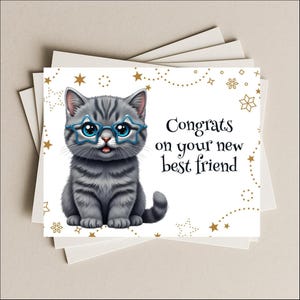 May include: Greeting card featuring a gray tabby cat wearing blue star-shaped glasses. The card reads "Congrats on your new best friend" in black text, with gold star and dot accents.