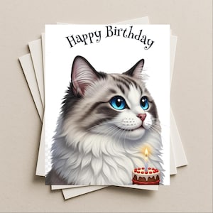 May include: A birthday card featuring a fluffy cat with blue eyes and a small birthday cake with a lit candle. The words "Happy Birthday" are written above the cat's head. The card is on a stack of white cards.