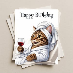 May include: A birthday card featuring a cartoon cat wearing a white robe and towel, holding a glass of red wine. The card reads "Happy Birthday" in black script. Several blank cards are stacked behind the main card.