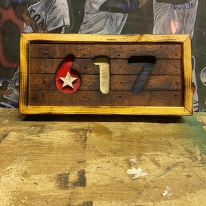 May include: Wooden sign with the numbers "617" cut out. The number 6 is red with a white star, 1 is beige, and 7 is blue. The sign is framed in a light brown wood.