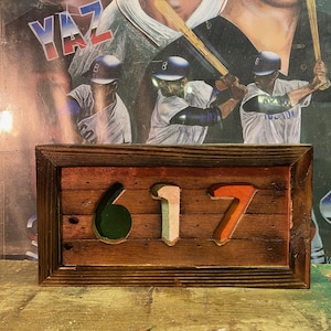 May include: Wooden sign with the numbers 617 cut out, painted in green, white, and orange. The sign is framed in dark brown wood and set against a baseball-themed background featuring baseball players and the word "YAZ".
