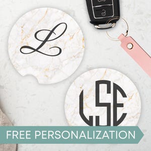May include: Two car coasters with a white marble design and gold veining. One coaster has a black cursive "L", the other has a black monogram. The text "FREE PERSONALIZATION" is at the bottom. A car key is in the background.