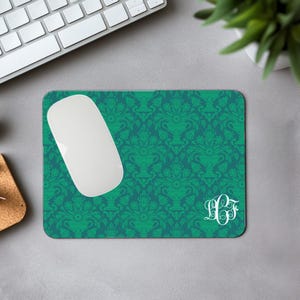 May include: A teal damask patterned mouse mat with a white monogram and a white wireless mouse. The mouse mat is on a grey surface, with a keyboard and plant in the background. The pattern features vase-like shapes.