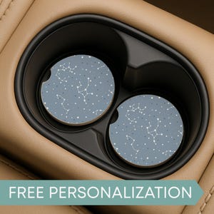 May include: Two round car coasters with a constellation and star pattern in white on a dusty blue background. The coasters are in a car cup holder. The text "FREE PERSONALIZATION" is at the bottom.