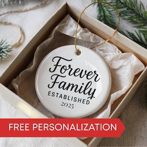 May include: A white ceramic ornament with the words "Forever Family ESTABLISHED 2025" in black script. The ornament has a gold string for hanging and is presented in a gift box. The bottom of the image has a red banner that says "FREE PERSONALIZATION".
