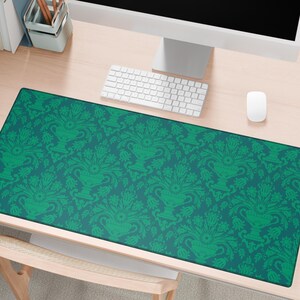 May include: A rectangular desk mat with a teal background and a repeating green floral pattern. The pattern features stylized vases and foliage. The mat is placed on a light-coloured wooden desk, with a keyboard, mouse, and monitor visible.