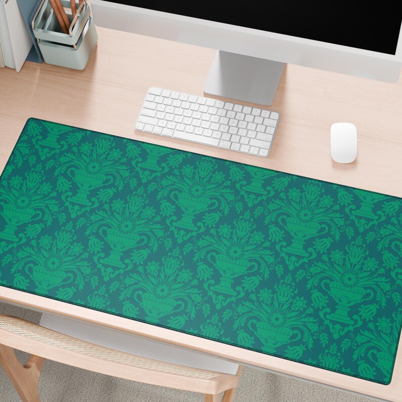 May include: A rectangular desk mat with a teal background and a repeating green floral pattern. The pattern features stylized vases and foliage. The mat is placed on a light-coloured wooden desk, with a keyboard, mouse, and monitor visible.