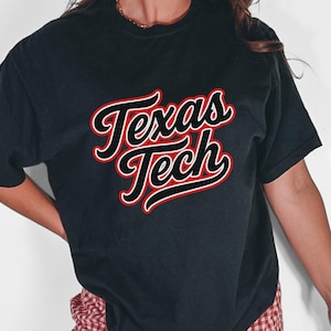 May include: Black t-shirt with the words "Texas Tech" in a red and white script design. The shirt is short-sleeved and appears to be made of a soft material. The text is outlined in red and white.