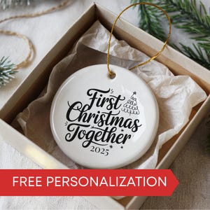 May include: A white ceramic Christmas ornament with the text "First Christmas Together 2025" in black script. The ornament is round and has a gold string for hanging. It is presented in a gift box with tissue paper.