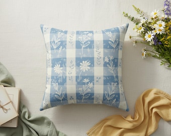 May include: Square throw pillow with a blue and white gingham pattern, adorned with floral and leaf illustrations. The pillow is surrounded by a bouquet of daisies and other flowers, with a yellow cloth and a stack of cards.