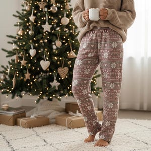 May include: Cozy holiday scene with a decorated Christmas tree and a person wearing patterned pajama pants. The pants feature a repeating design of snowflakes, hearts, and other festive shapes in shades of pink, gray, and white. A white mug is held.