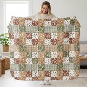 Pode incluir: Um edredão patchwork com quadrados de tecidos florais, xadrez e lisos em tons de castanho, verde e branco. O edredão é segurado, mostrando o seu design. É um elemento de decoração de casa acolhedor.