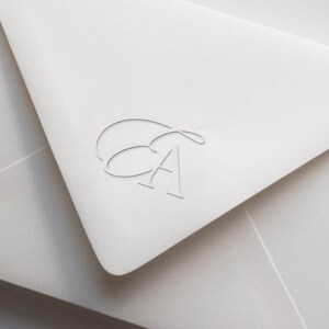 Può includere: Primo piano di una busta bianca con un monogramma. Il monogramma presenta le lettere "E" e "A" in un delicato carattere corsivo. La busta è realizzata in carta strutturata.