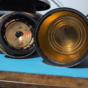 May include: Two open, empty tin cans with gold-colored interiors and black rims. One can has black residue on the inside. The cans are on a blue surface, with a blurred background.