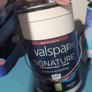 May include: A cylindrical gallon can of Valspar Signature high-hiding paint and primer with an eggshell interior finish. The dark blue label features white text, a metal handle, and a white ribbed plastic lid. The text reads Valspar Signature.