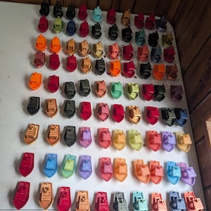 May include: A large collection of small, 3D-printed plastic toy boats arranged in rows. The boats feature a variety of solid colors including red, orange, yellow, green, blue, purple, black, and brown. Each boat has a cabin and a hull design.
