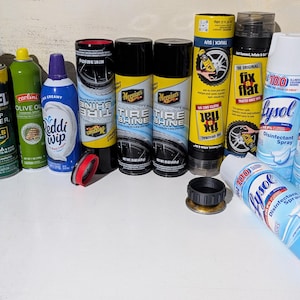 Puede incluir: Una colección de varios botes de aerosol que incluyen Repel Camp Fogger, aceite de oliva Carlini, Reddi-wip, Meguiar's Tire Shine, Fix-A-Flat y tres botes de spray desinfectante Lysol, con dos tapas de plástico negro y rojo sobre la superficie.