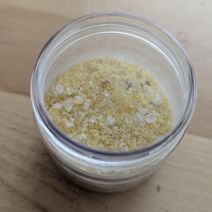 Peut inclure: Un pot en verre transparent rempli de granules cristallins jaune clair. Les granules ont un aspect légèrement translucide et de tailles variables. Le pot est posé sur une surface en bois brun clair.