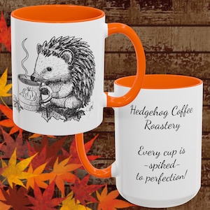May include: White ceramic coffee mug with an orange interior and handle. The mug features a black and white illustration of a hedgehog drinking coffee from a mug with a pumpkin design. The text on the mug reads "Hedgehog Coffee Roastery" and "Every cup is -spiked- to perfection!"