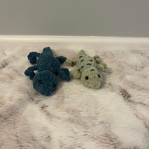 May include: Two crocheted alligator plush toys. One is teal, the other is light green with dark green spots. Both have small black eyes and are lying on a fluffy, light pink and white surface.