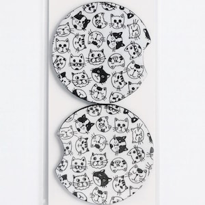 May include: Two white car coasters with a black cat face pattern. The coasters have a circular shape with a curved notch on one side. The cat faces are in various poses and expressions.