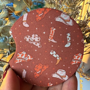 May include: A round, rust-colored car coaster featuring a repeating pattern of cowboy hats and boots in shades of orange and white. The coaster is designed to absorb moisture and protect car surfaces.
