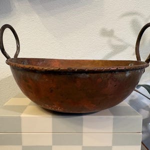 May include: A copper bowl with two arched handles. The bowl has a wide, shallow shape with a textured rim. The copper has a warm, reddish-brown color with areas of darker patina, suggesting age and use. The bowl is likely for decorative or serving purposes.