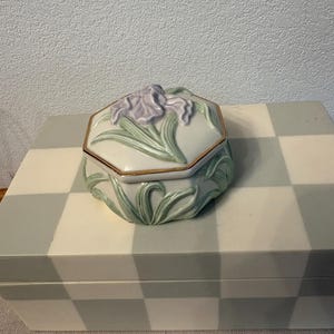 May include: A ceramic jewelry box with a hexagonal shape and a lid featuring a lavender iris flower. The box is white with green leaves and gold trim. The box is sitting on a white and gray checkered surface.