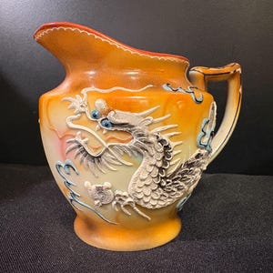 May include: A ceramic pitcher featuring a gradient orange and cream glaze with a prominent, raised relief of a dragon. The dragon has textured scales, a long body, and blue-painted eyes. The pitcher includes a curved handle and a wide pouring spout.