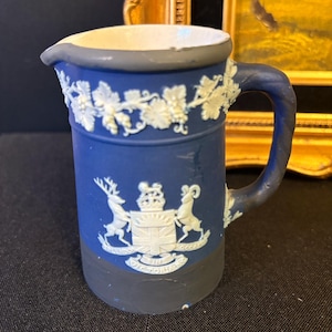 May include: A blue ceramic pitcher featuring white relief decorations of grapevines around the rim and a central coat of arms with a crown, a shield, and two animals. The base and rim are dark gray, and the handle is blue with a twisted texture.
