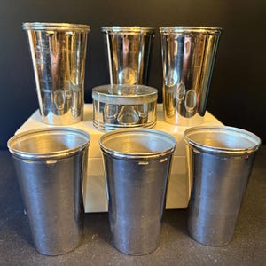 May include: A set of five silver-toned metal tumblers with a smooth, polished finish and a single small, round, lidded metal container positioned in the center. The tumblers are arranged in two rows, with three in the front and two in the back.