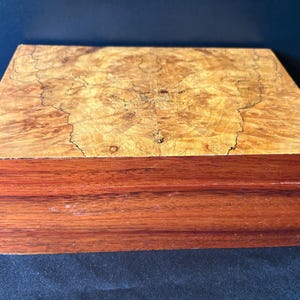 May include: A rectangular wooden box featuring a light tan burl wood lid with a complex, swirling grain pattern and dark brown, straight-grained wood sides. The box has a flat top and a horizontal seam indicating a hinged or removable lid design.