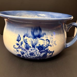 May include: A blue and white porcelain chamber pot with a handle. The pot features a floral design in shades of blue against a white background. The rim and handle are a darker blue, creating a classic, vintage aesthetic.