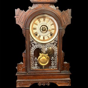 May include: A dark brown wooden mantel clock featuring an arched top, a circular clock face with Roman numerals, and a glass panel below displaying a gold-colored pendulum. The clock case has intricate carved details and a rectangular base.