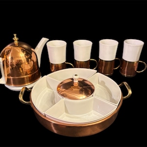 May include: A tea set featuring a teapot with a copper dome and white handle, four white mugs with copper bases, and a round serving dish with a copper-trimmed lid and five white interior compartments, all set against a black background.