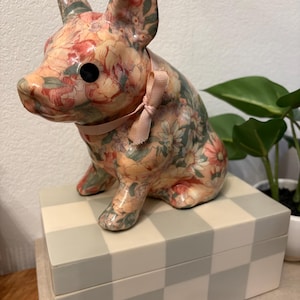 May include: A ceramic pig figurine with a floral pattern in shades of pink, peach, and green, wearing a pink ribbon. The pig sits atop a gray and white checkered box. Decorative home decor.